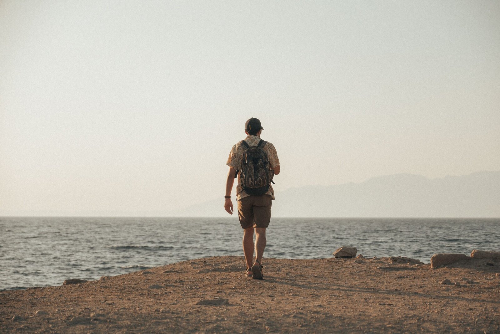Home A lone traveler with a backpack walks towards the distant horizon along a tranquil ocean shore.