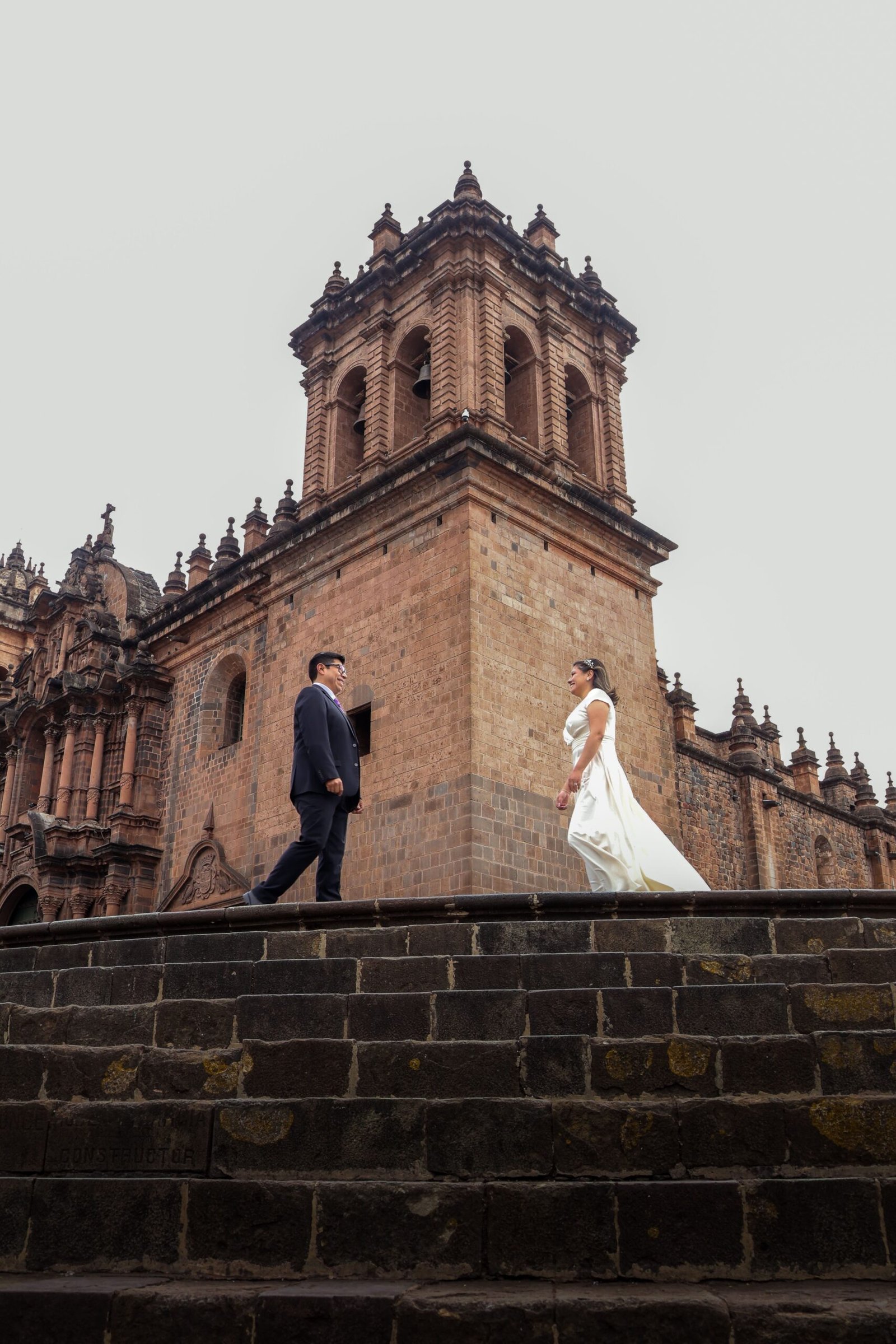 Home A bride and groom stand romantically on stone steps, with historic church architecture in the background.